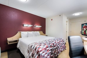 A hotel room with a bed, a desk with a chair, a wall-mounted light fixture, and a door.