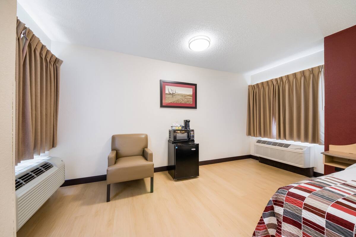 A hotel room with a bed, a chair, a small table with a coffee machine, an air conditioner, and a framed picture on the wall.