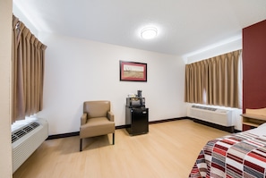 A hotel room with a bed, a chair, a small table with a coffee machine, an air conditioner, and a framed picture on the wall.