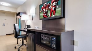 Quarto de hotel com televisão de tela plana na parede, escrivaninha com computador, micro-ondas e cadeira de escritório preta.