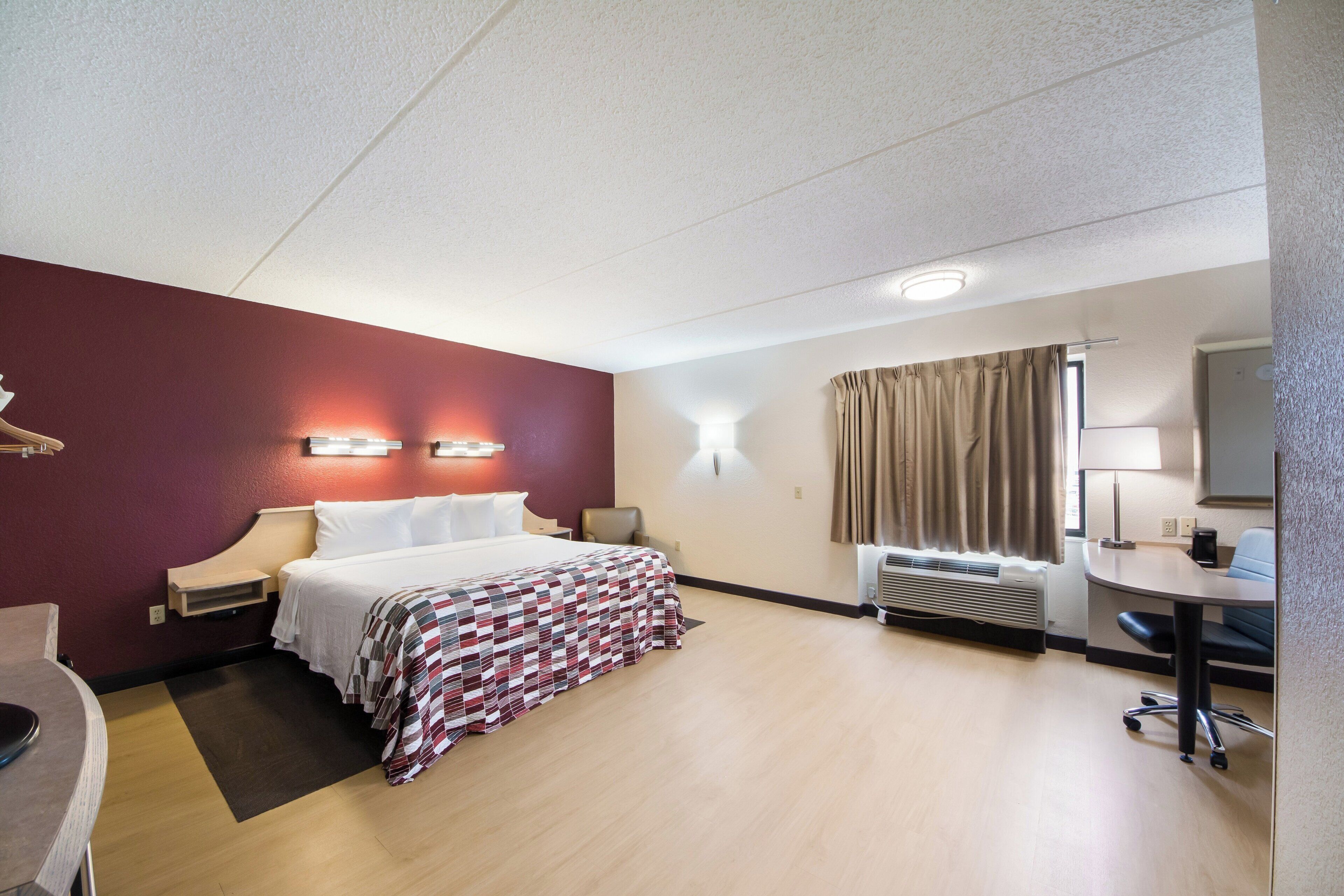 A hotel room with a bed, bedside tables, a desk, a chair, a desk lamp, a heater, and a window with curtains.