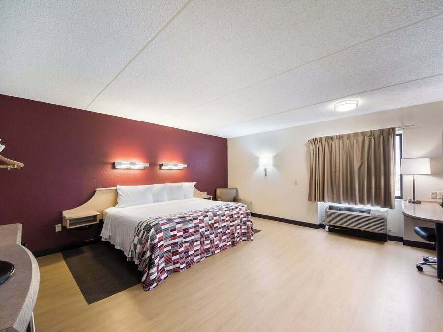 Habitación de hotel con cama, mesitas de noche, escritorio, silla, lámpara de escritorio, calefactor y ventana con cortinas.