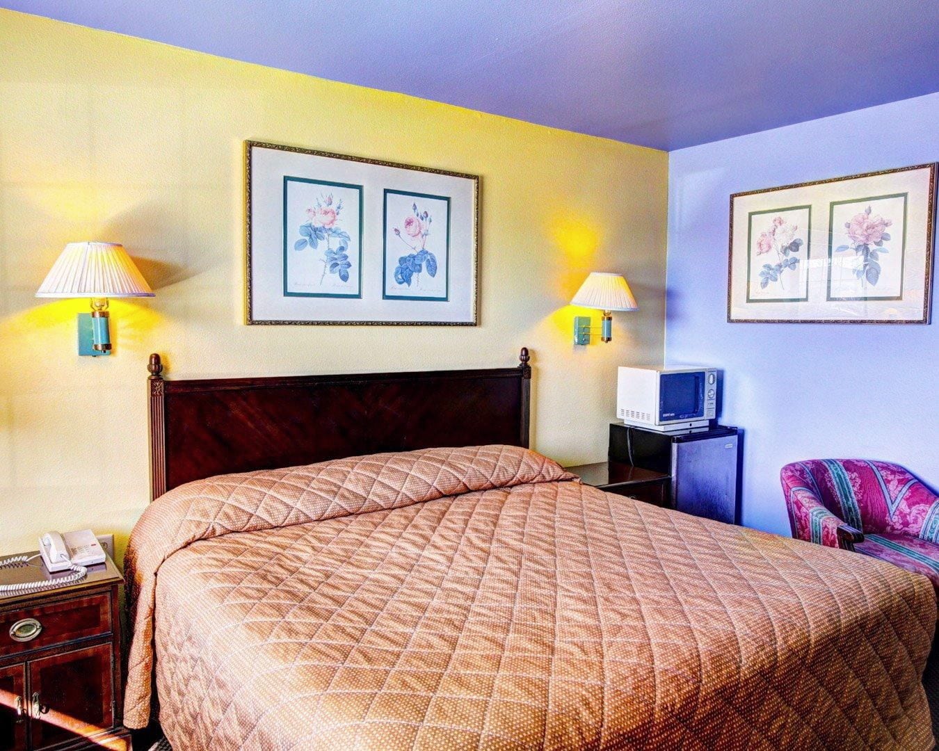 A hotel room with a large bed, two wall-mounted lamps, a television on a stand, and two framed floral artworks on the wall.