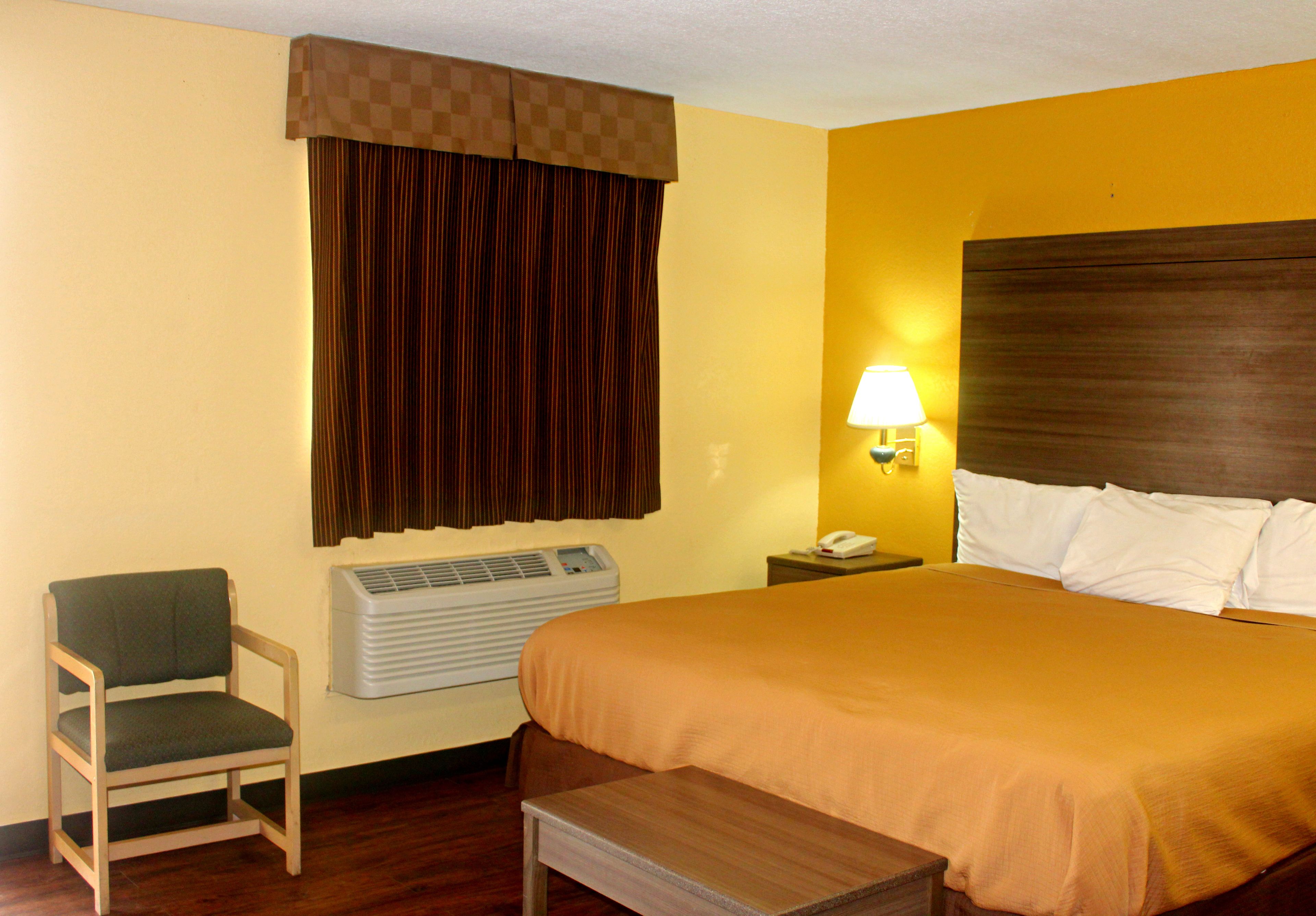 Una habitación de hotel con cama, una silla, mesita de noche, lámpara y aire acondicionado.