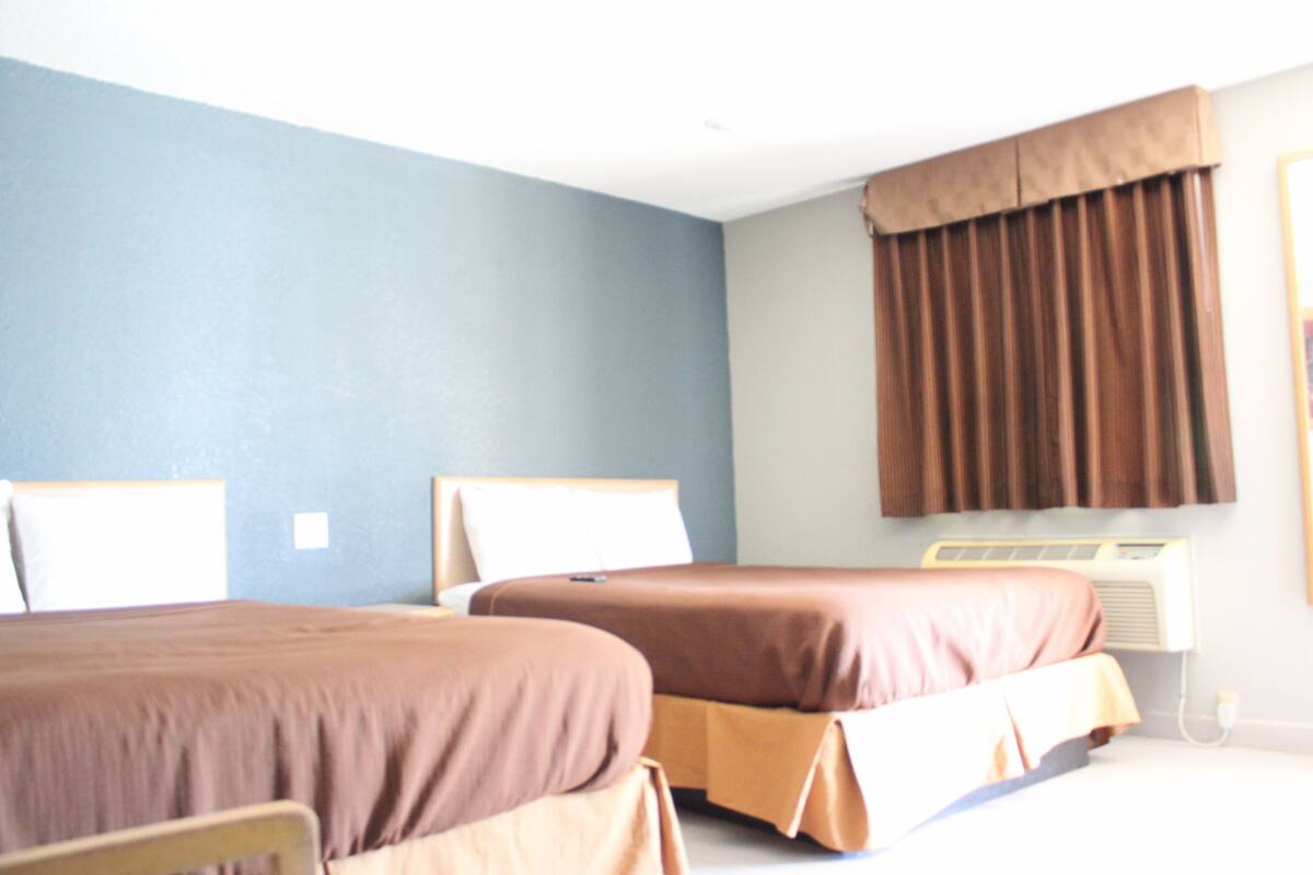 A hotel room with two beds, a window with curtains, and an air conditioning unit.