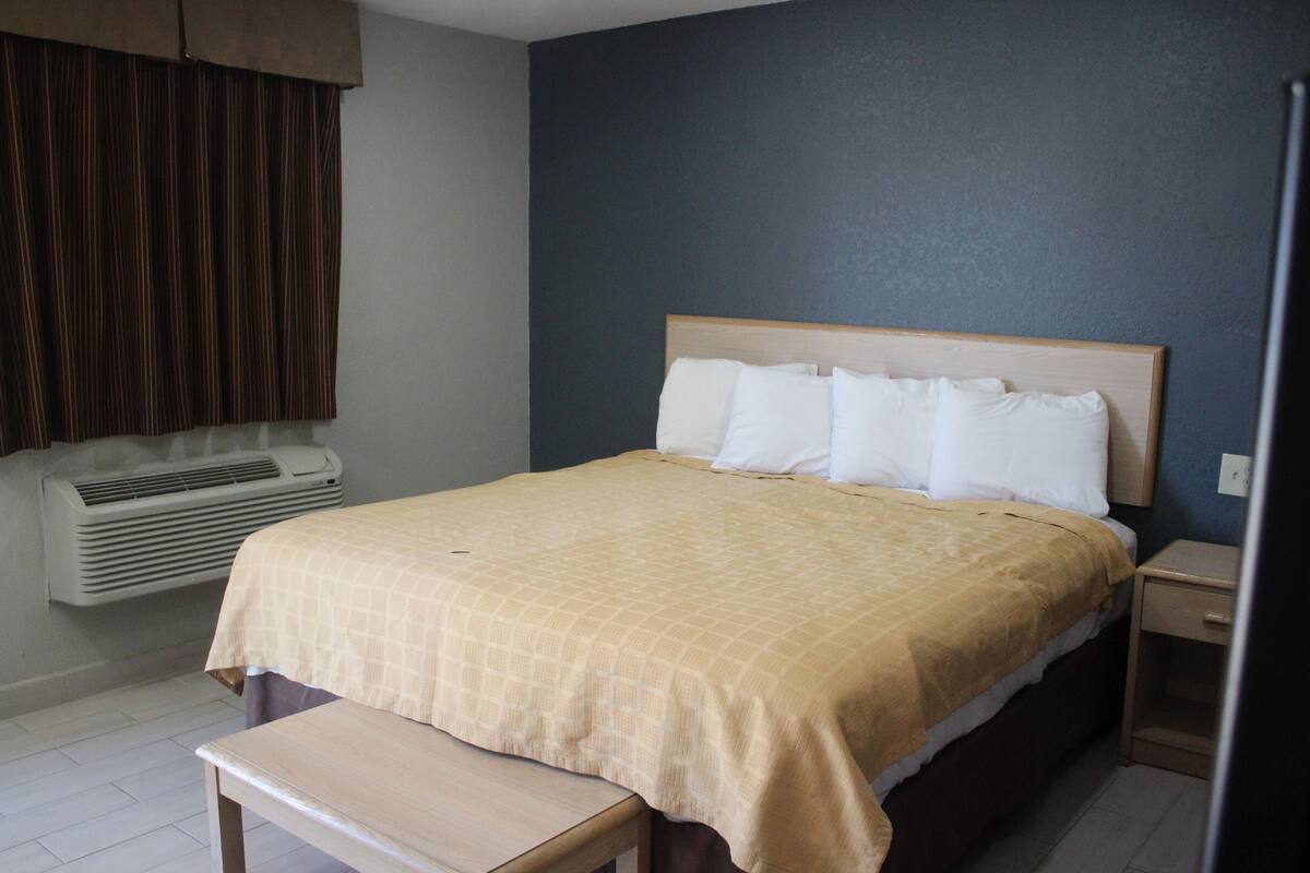 A neatly made bed with a beige bedspread and white pillows, a bedside table, a bench, and a wall-mounted air conditioner.