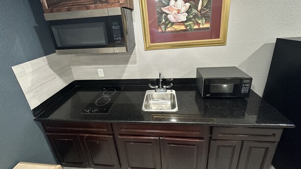A kitchen with dark wooden cabinets, a black countertop, a stainless steel sink, and two black microwaves. - Village Inn Cotulla (Cotulla)