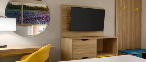 A hotel room with a bed, a desk with a chair, a television, a wardrobe, and a mirror with a floral view.