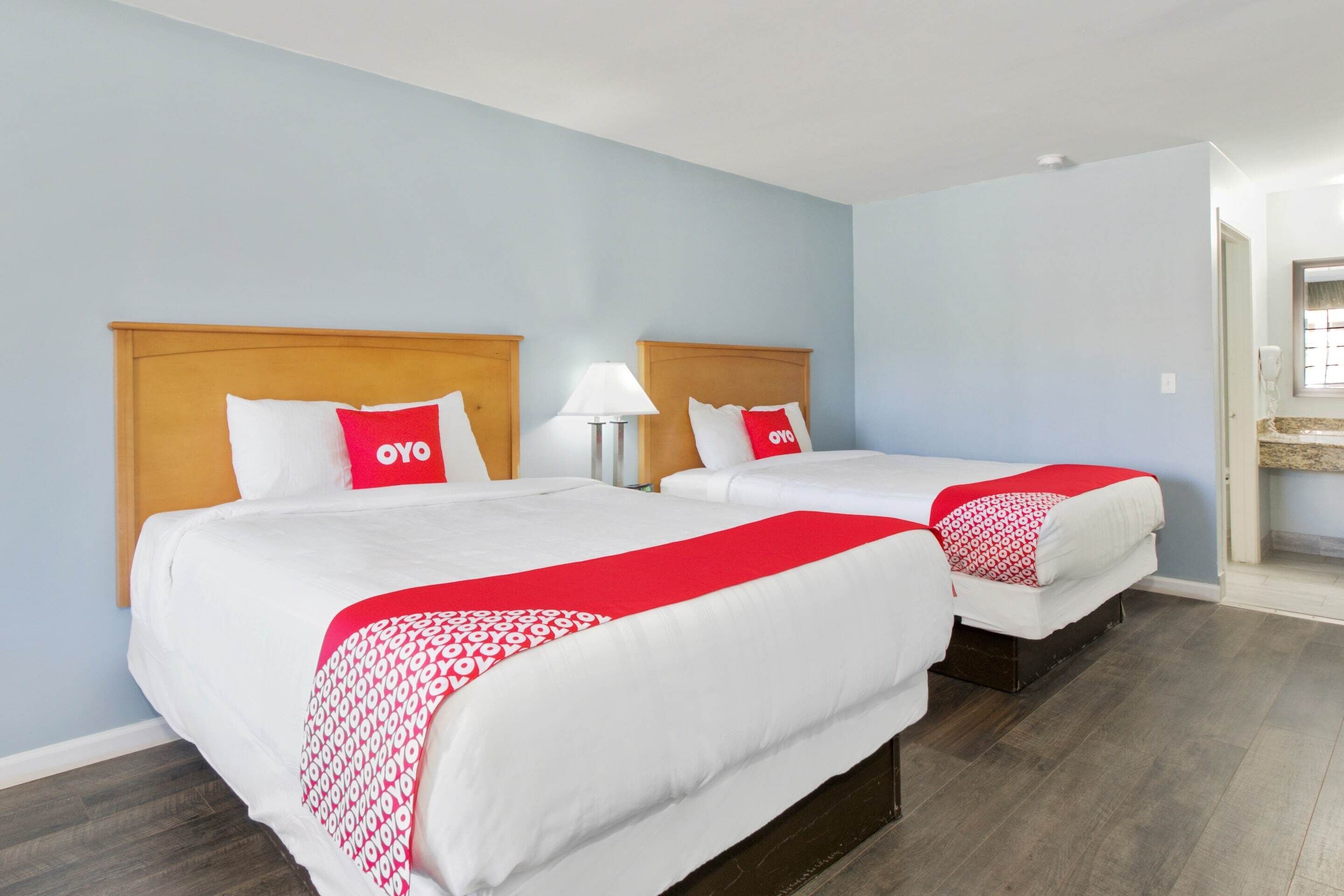 A hotel room with two beds, each with a wooden headboard, white bedding, and red accents. There is a nightstand with a lamp between the beds.