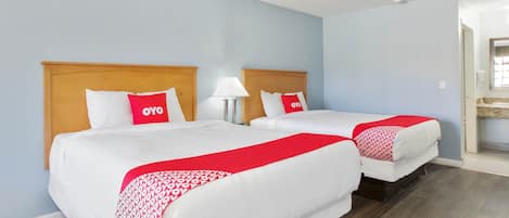 A hotel room with two beds, each with a wooden headboard, white bedding, and red accents. There is a nightstand with a lamp between the beds.