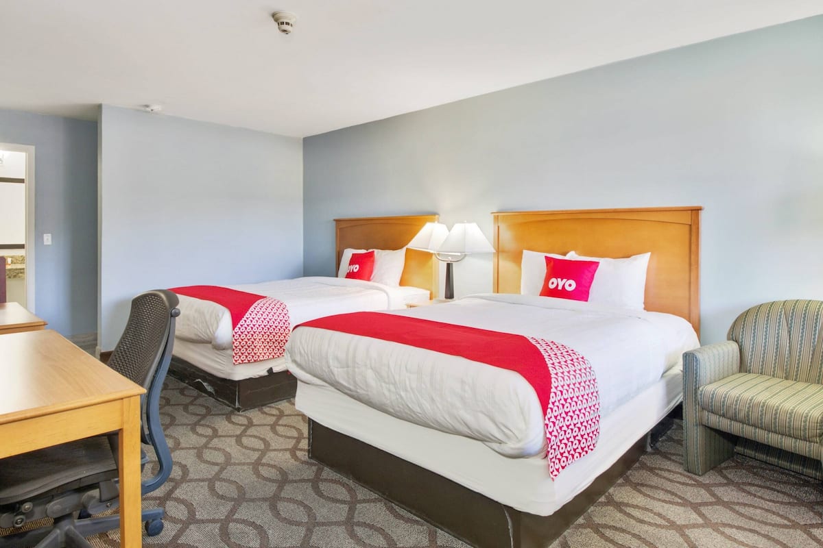 A hotel room with two beds, each with a red and white blanket, a wooden headboard, and a lamp. There is a desk with a chair and a patterned carpet.