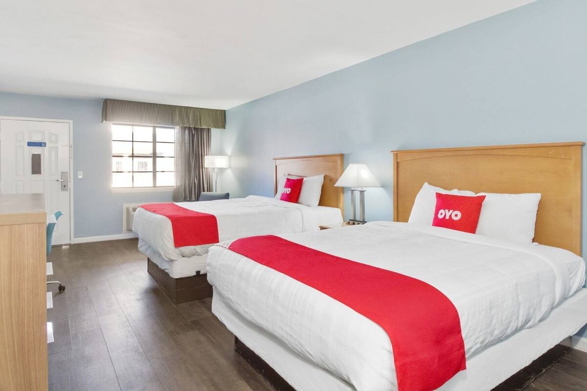 A hotel room with two beds, a desk, a chair, a window with curtains, and a door.