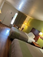 Habitación de hotel con dos camas, suelo de madera, pared verde decorativa, una puerta con manija y una ventana con cortinas.