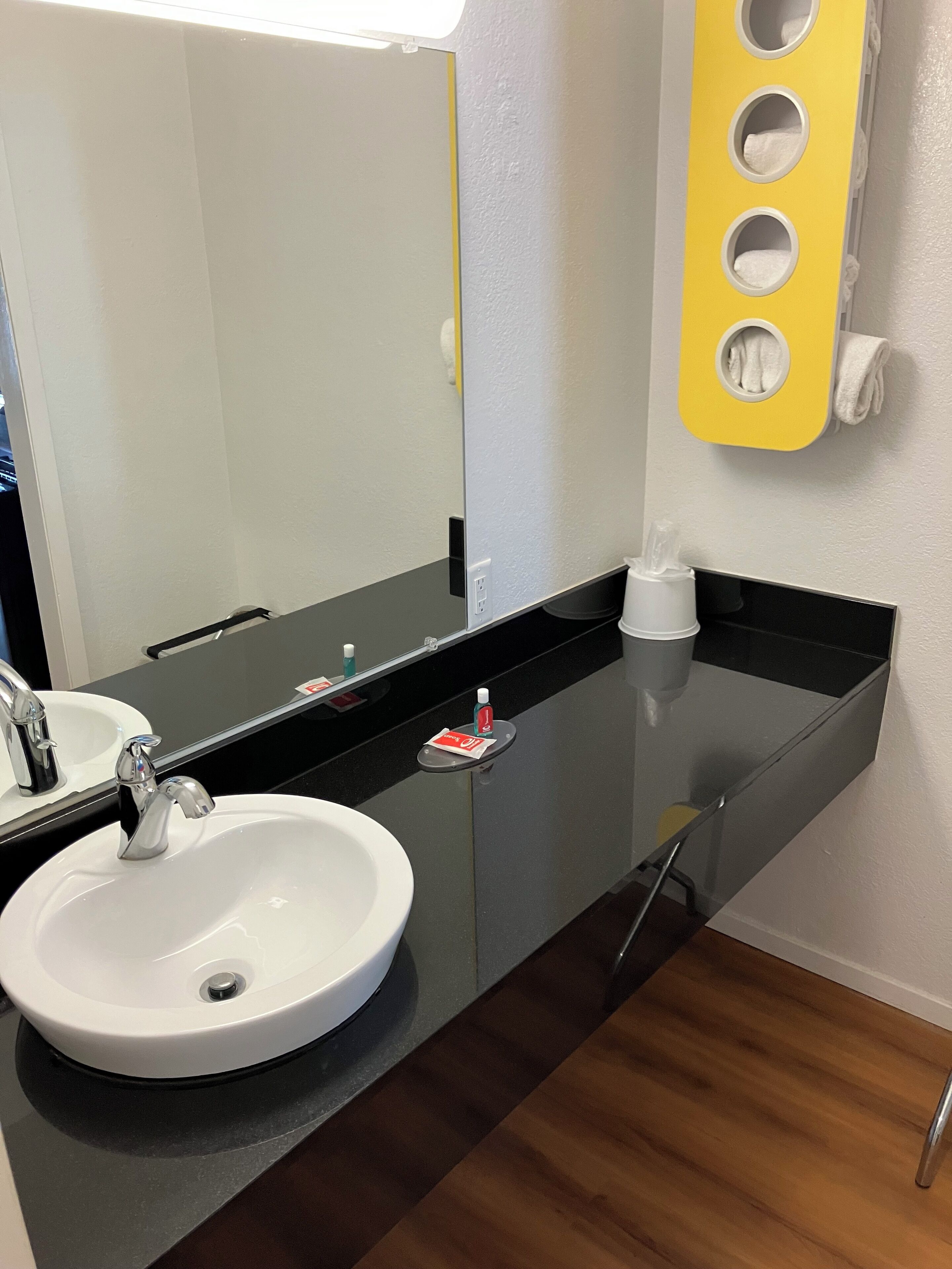 A bathroom with a white sink, black countertop, and a mirror. There is a yellow towel rack with three towels, a white soap dispenser, and a small red and white sign on the countertop.