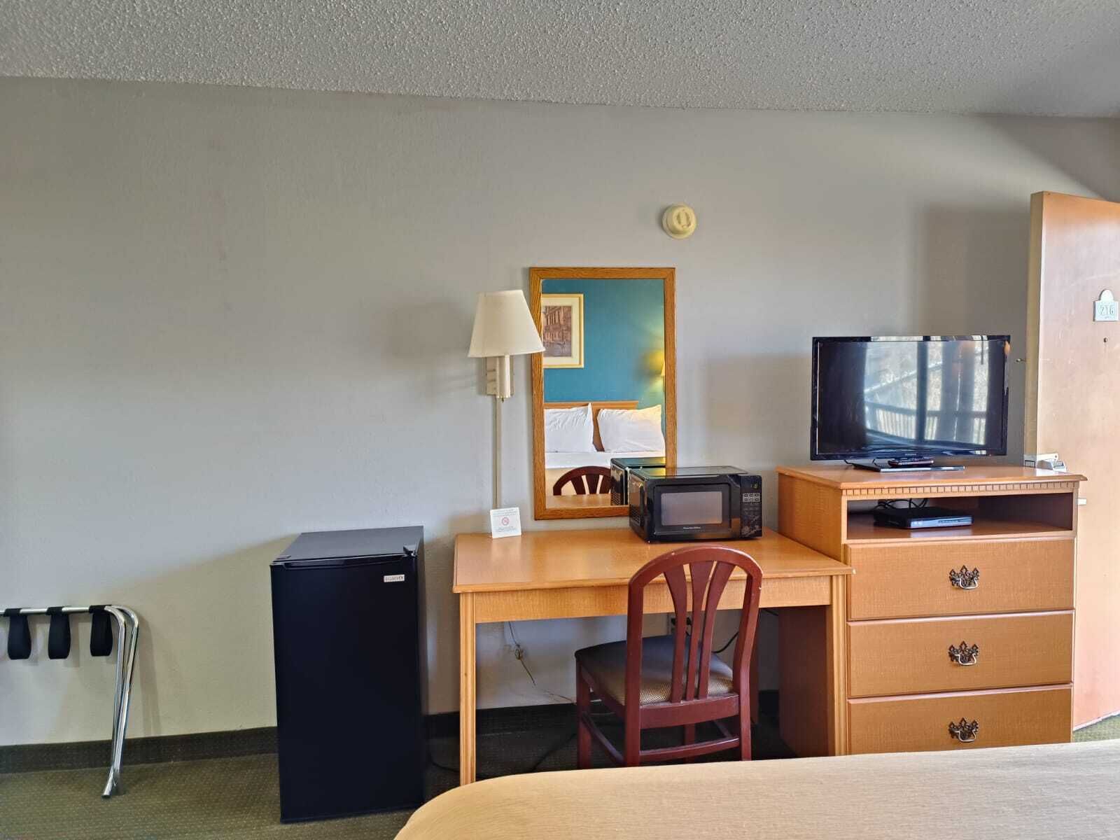 A hotel room with a bed, a desk with a computer, a television, a microwave, a chair, a small refrigerator, and a bedside table with a lamp.