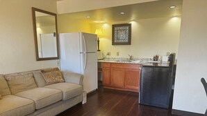 A compact living space with a sofa, a refrigerator, a kitchen area with wooden cabinets, and a small black refrigerator. - Super 8 by Wyndham Giddings (Giddings)