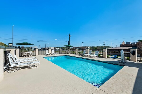 A rectangular outdoor swimming pool with lounge chairs and umbrellas, surrounded by a concrete deck and a fence.