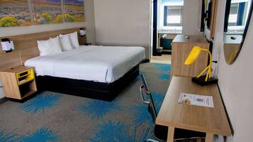 A hotel room with a bed, a desk with a lamp, a small shelf, and a painting on the wall.