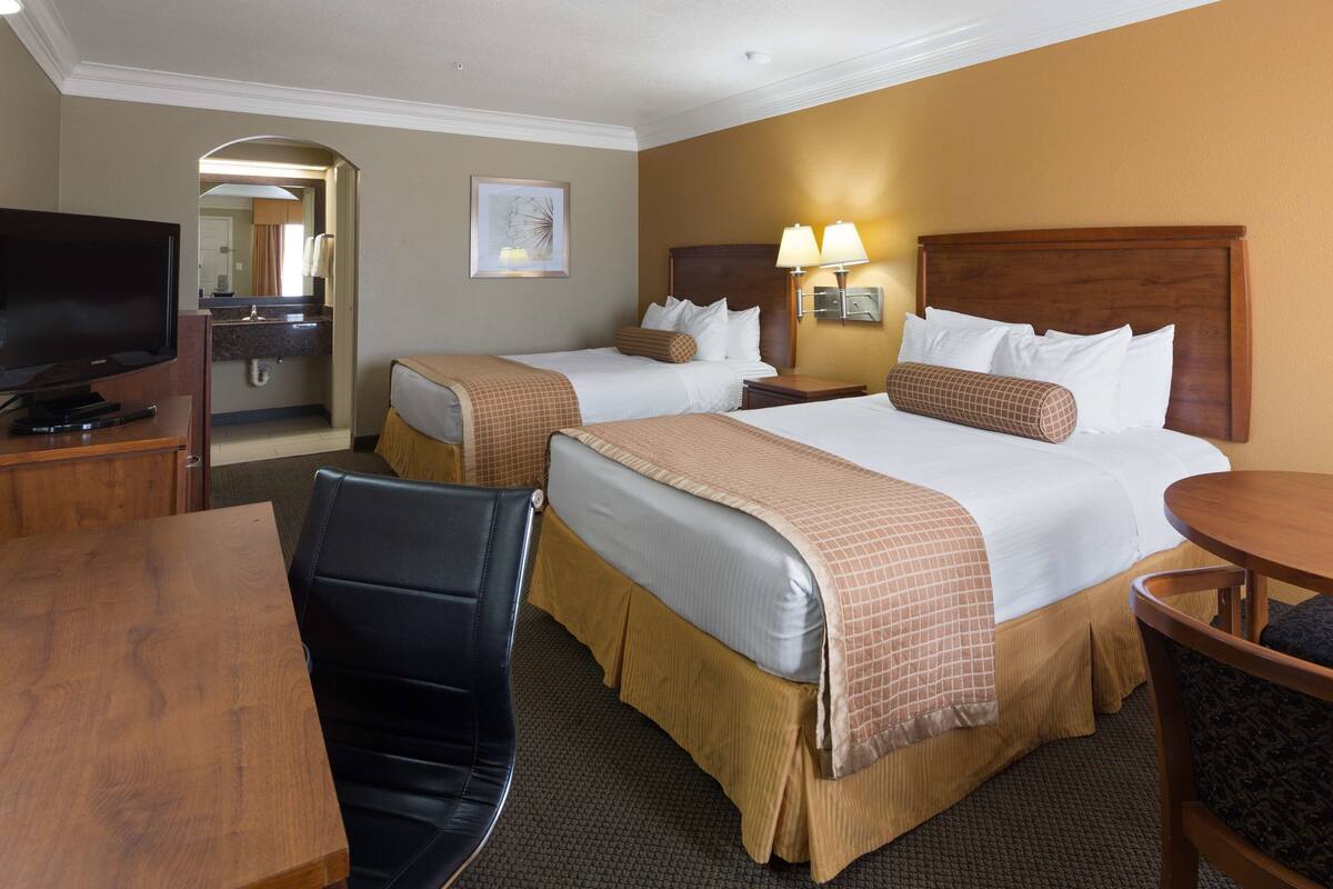 Quarto de hotel com duas camas, uma escrivaninha, uma cadeira, uma televisão e um quadro na parede.