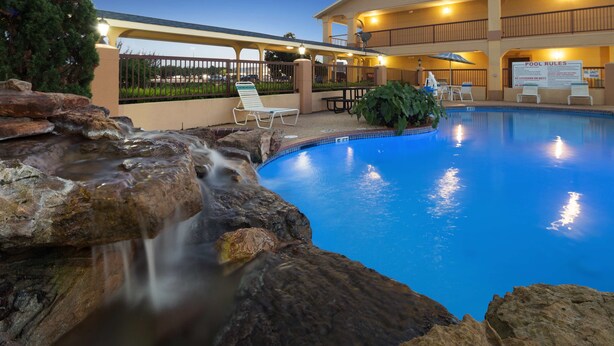 Una piscina con cascada, sillas de descanso y un cartel con las normas de la piscina.