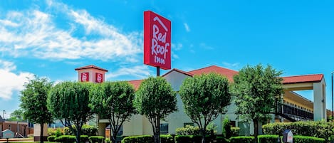 Un Red Roof Inn con un letrero rojo llamativo, rodeado de vegetación cuidada y un cielo azul despejado.