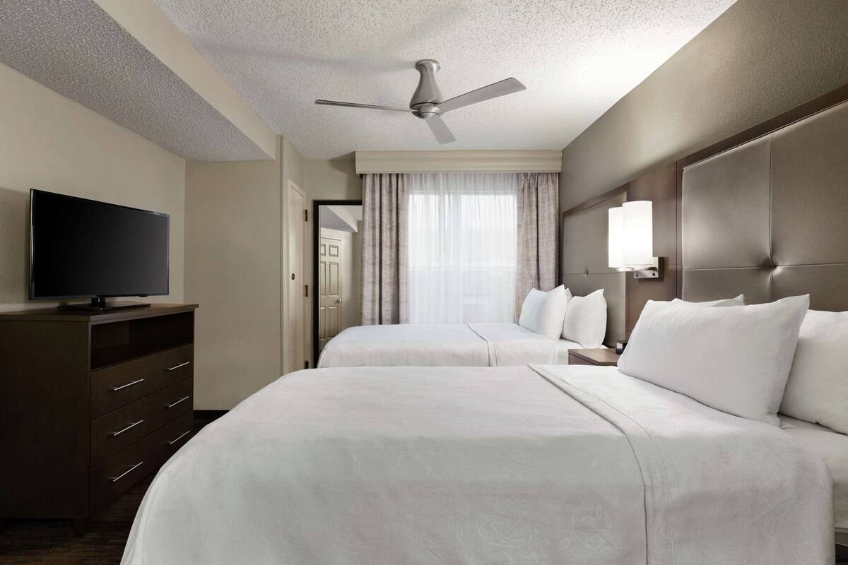 Hotellihuone, jossa on kaksi sänkyä, taulutelevisio, kattotuuletin ja suuri ikkuna, jossa on verhot.