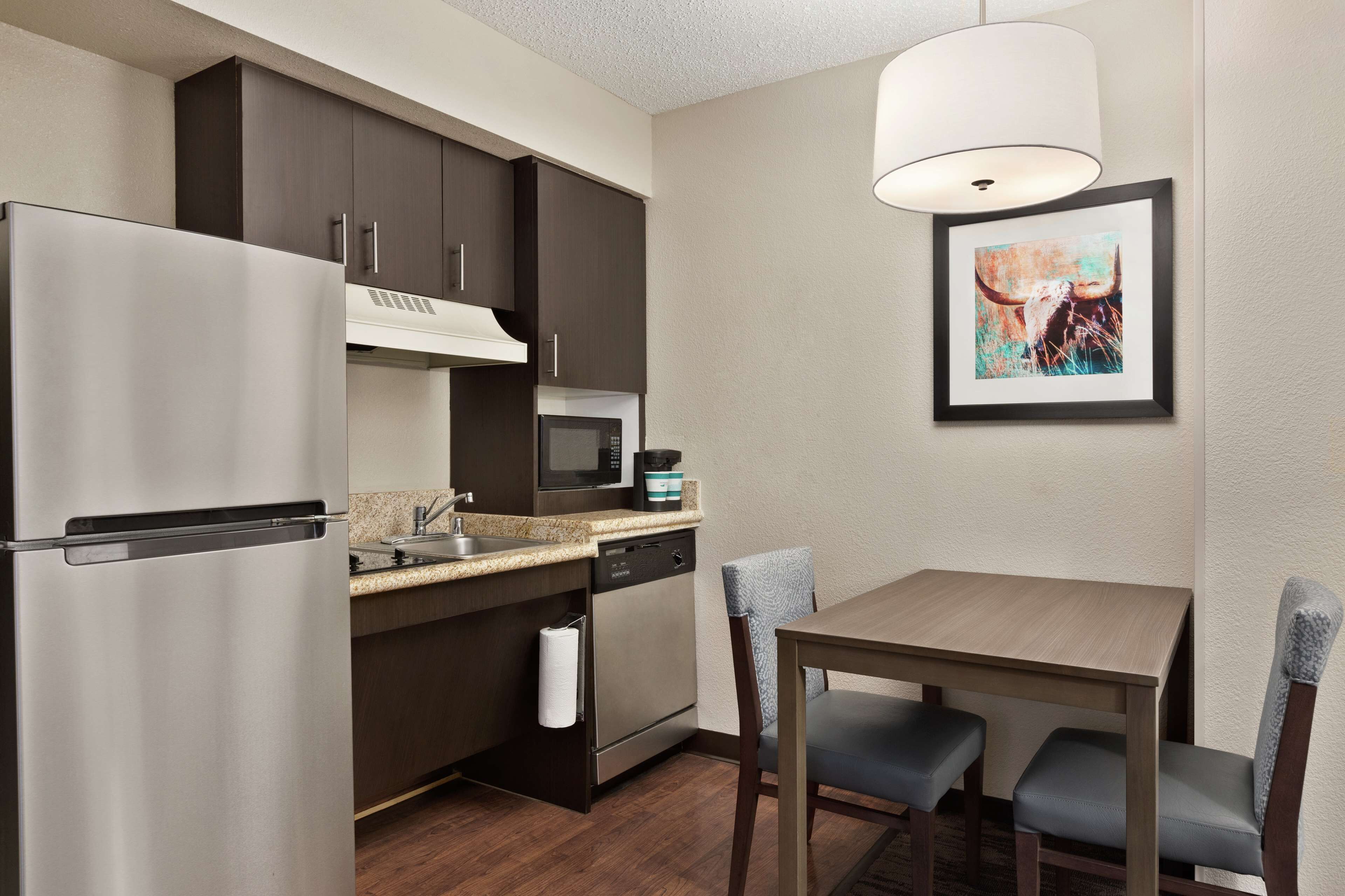 Cocina compacta con refrigerador, microondas y lavavajillas, una mesa de comedor con sillas y un cuadro en la pared.