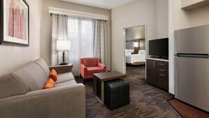A hotel room with a sofa, a chair, a small table, a TV, a refrigerator, and a bed. - Homewood Suites by Hilton North Dallas-Plano (Plano)