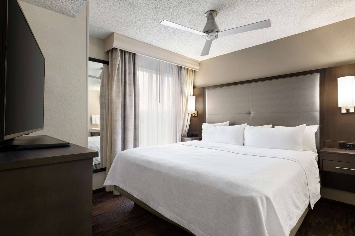Hotellihuone, jossa on suuri sänky, taulutelevisio, kattotuuletin ja yöpöytä, jossa on lamppu.