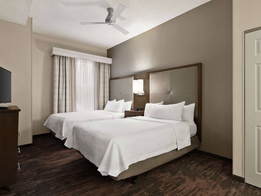 Habitación de hotel con dos camas, televisión, ventilador de techo y un ventanal grande con cortinas.
