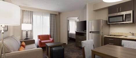 Habitación de hotel con sofá, sillón, reposapiés, mesa de comedor, kitchenette y televisor.