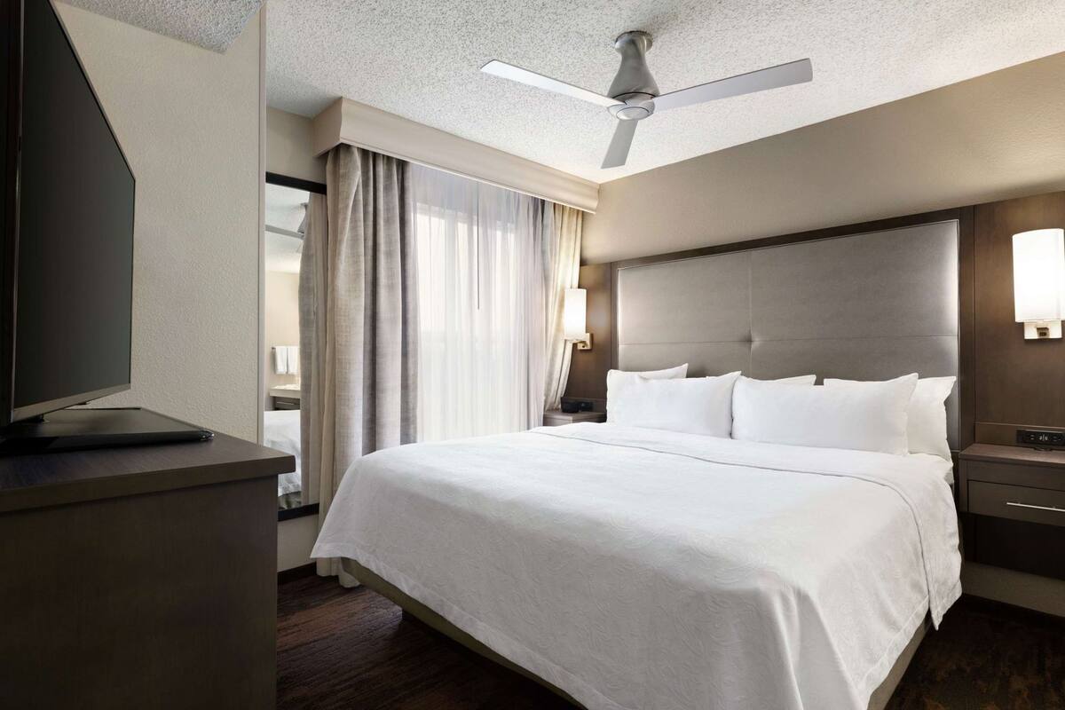 Hotellihuone, jossa on sänky, televisio, kattotuuletin ja yöpöytä, jossa on lamppu.