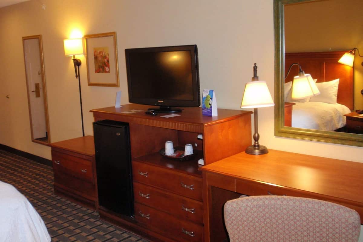 Une chambre d’hôtel avec un bureau en bois, une télévision, un miroir, une lampe, un lit et une chaise à motifs.