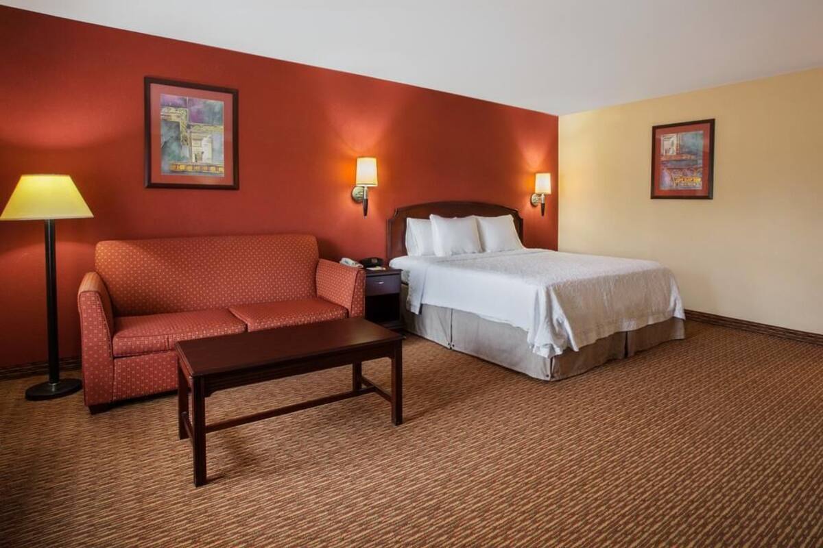 A hotel room with a red sofa, a bed with white bedding, a nightstand, a lamp, and two wall-mounted lights.