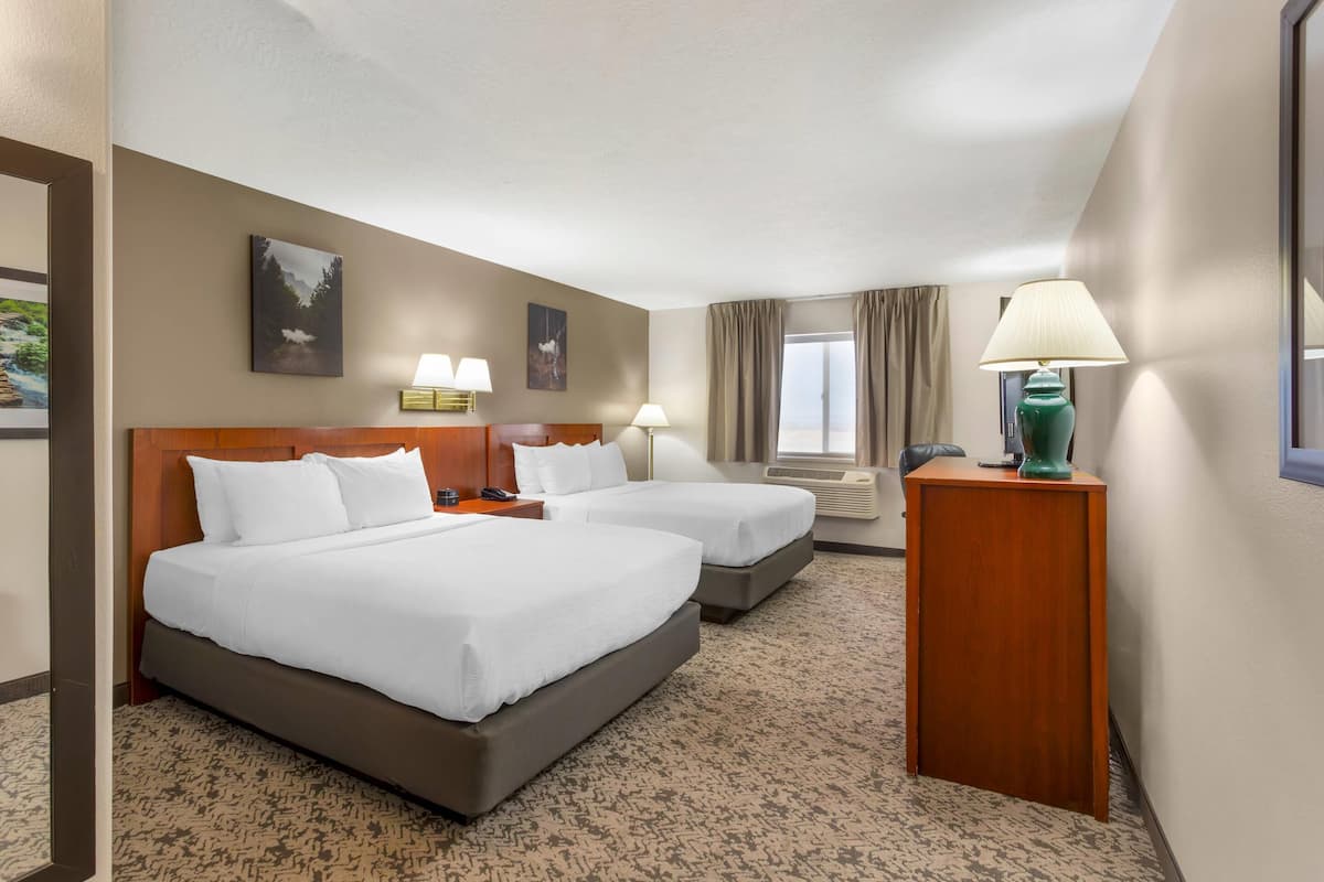 A hotel room with two beds, a wooden headboard, a desk with a lamp, and a window with curtains.