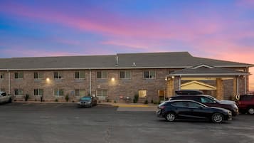 A multi-story hotel with a brick facade, a covered entrance, and a parking lot with several cars.