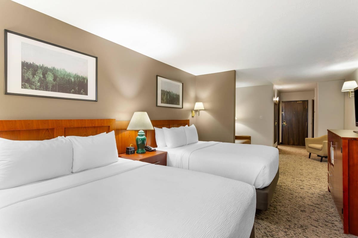 A hotel room with two beds, a wooden headboard, a desk with a lamp, and framed pictures on the wall.