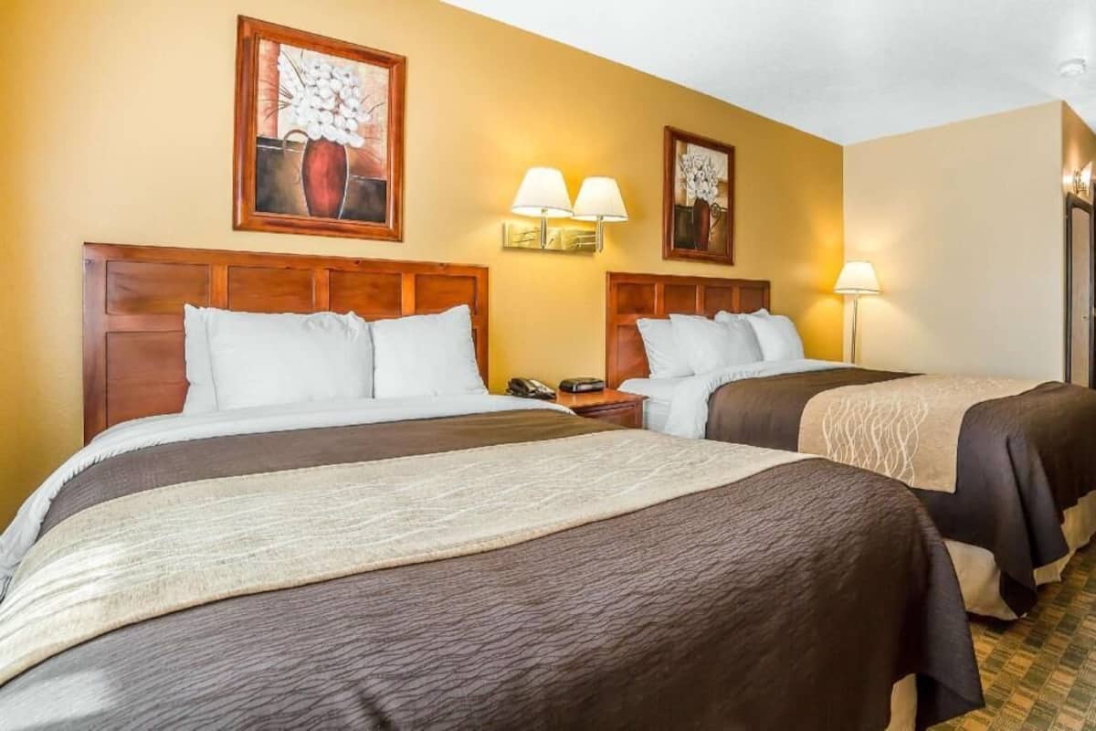 A hotel room with two beds, wooden headboards, wall-mounted lamps, and artwork.