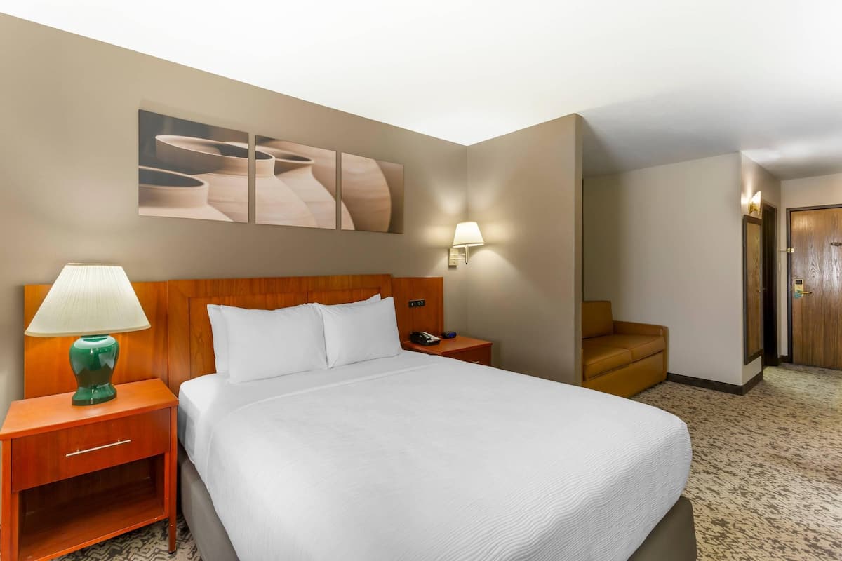 A hotel room with a large bed, a wooden headboard, a nightstand with a lamp, a wall-mounted phone, and a door leading to another room.