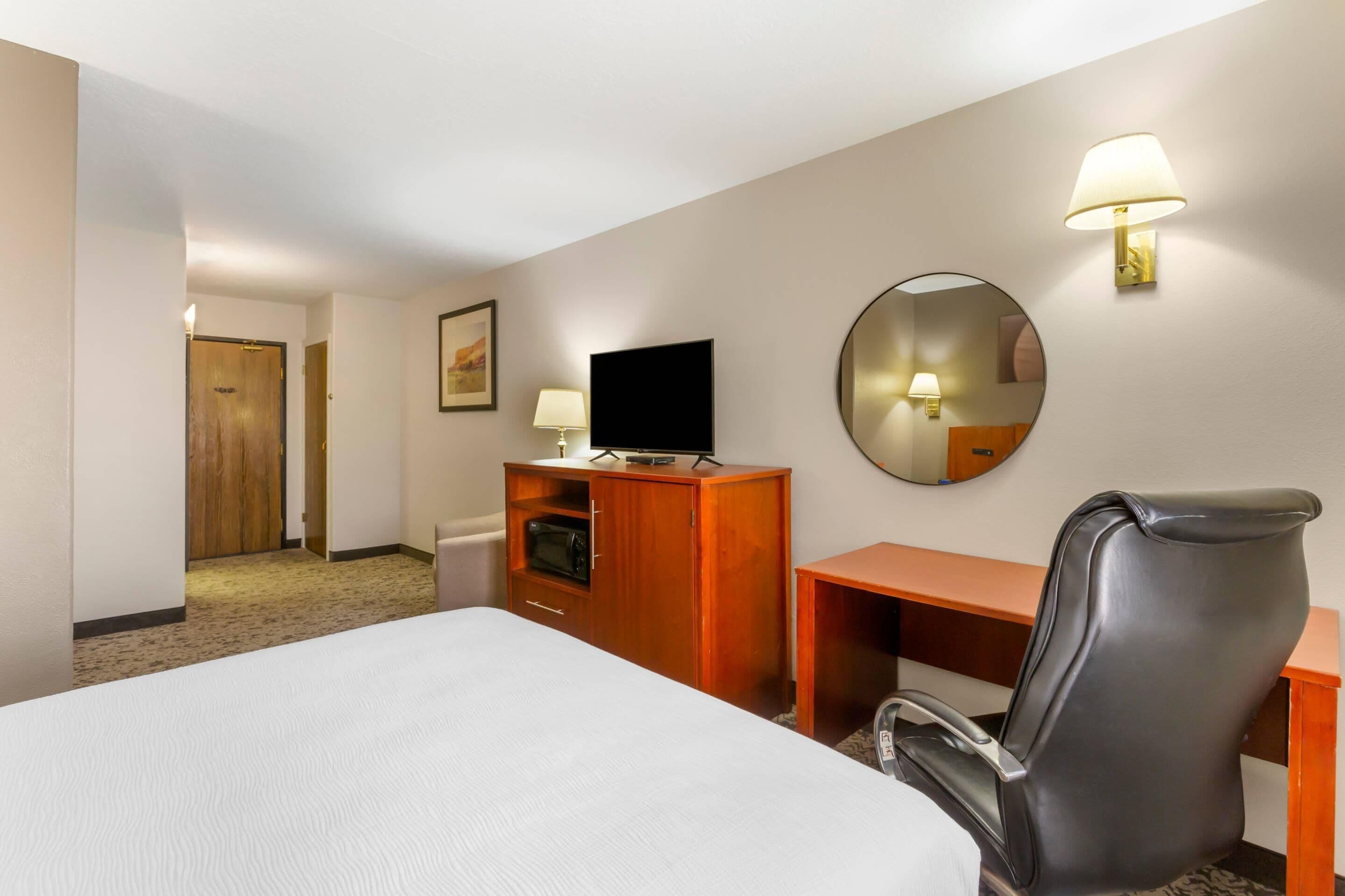 Habitación de hotel con cama, escritorio, silla, televisor, espejo y puerta que da a otra habitación.
