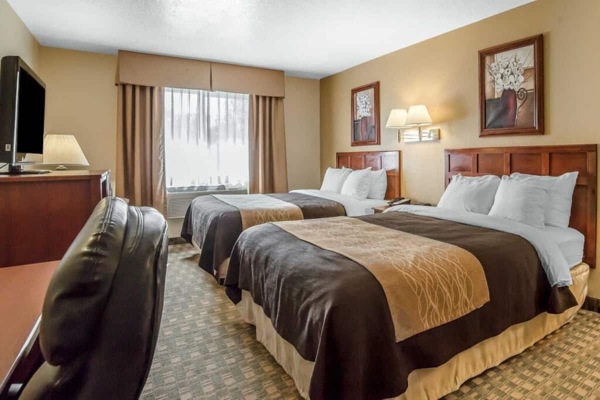 A hotel room with two beds, a desk, a TV, and a window with curtains.