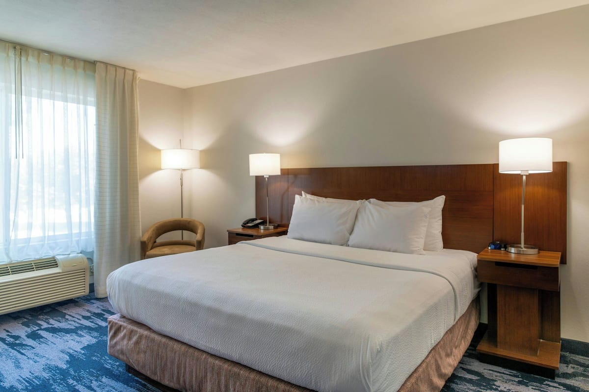 Una camera d'albergo con un letto grande, due comodini con lampade, una sedia, una scrivania con un telefono e una finestra con tende.
