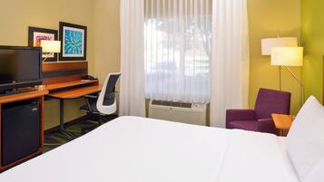 A hotel room with a bed, a desk with a TV, a chair, a purple armchair, a lamp, and a window with curtains.