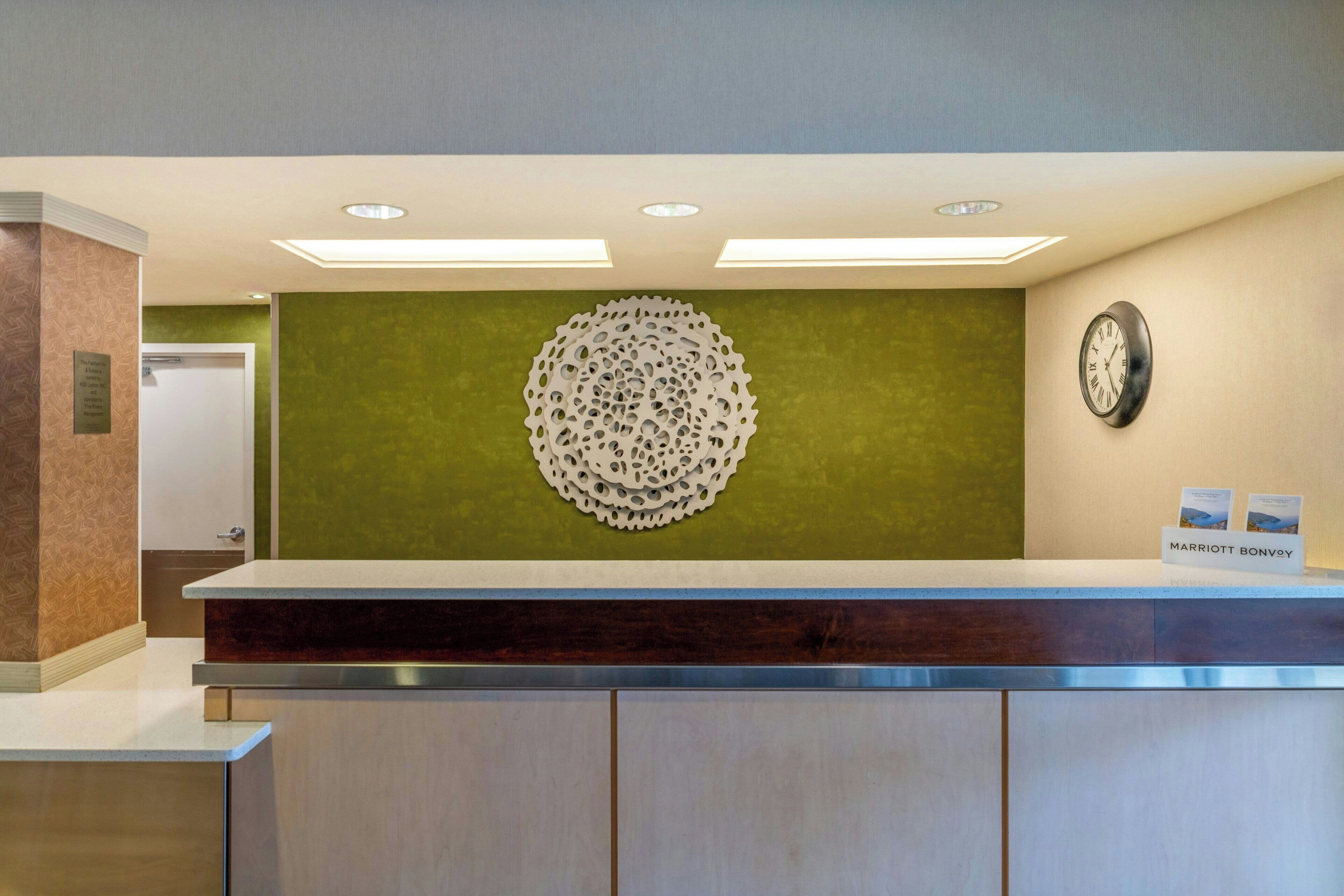 Un área de recepción con diseño moderno, que incluye un mostrador de recepción, un elemento decorativo en la pared, un reloj y un cartel con el nombre del hotel.
