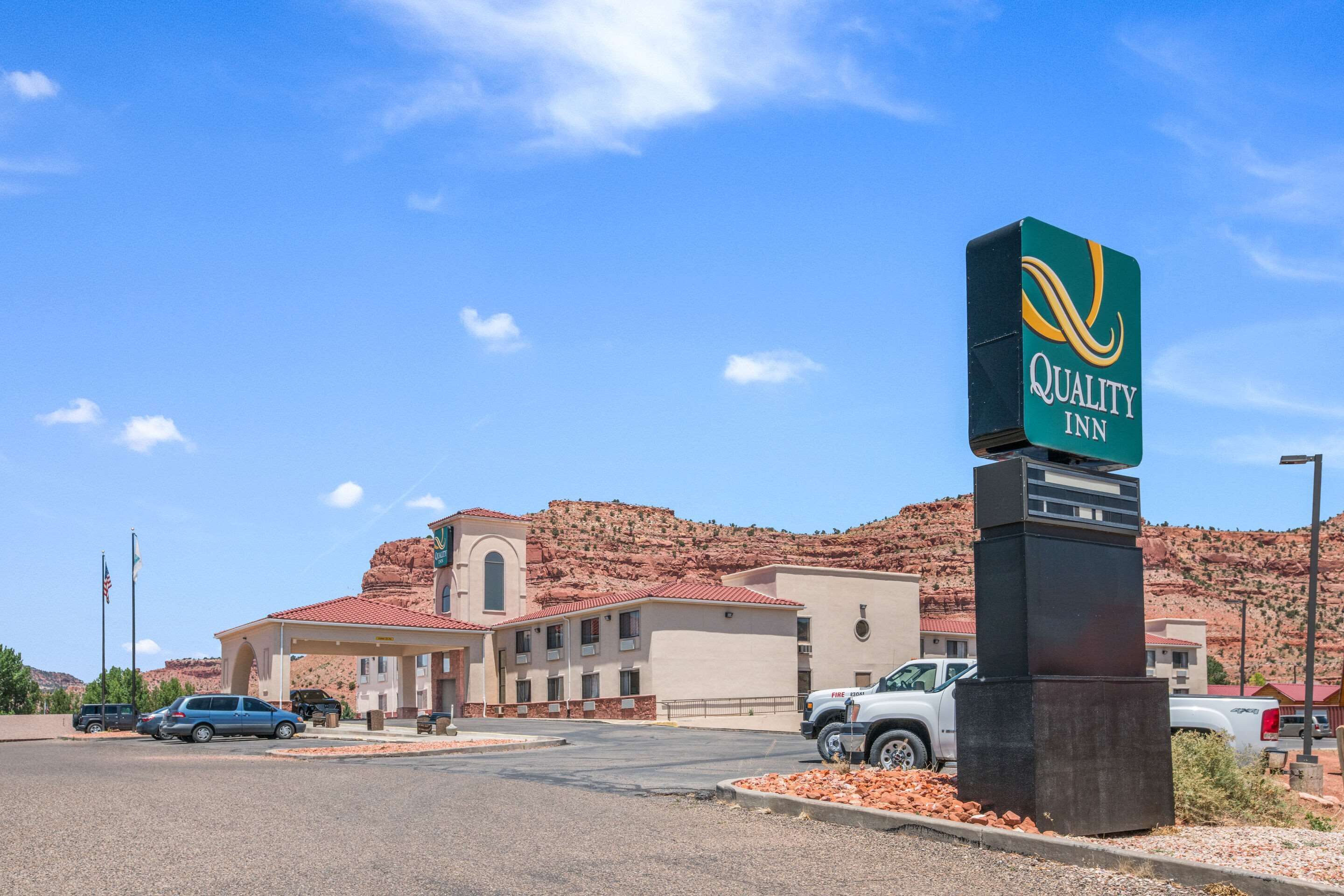 Señal de Quality Inn con montañas al fondo, un edificio de techo rojo y un estacionamiento con vehículos.
