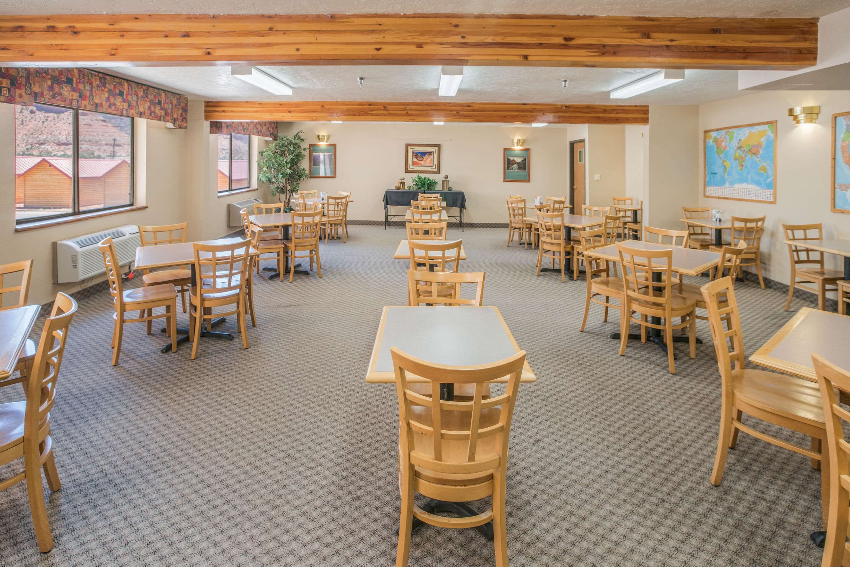 A spacious dining area with multiple wooden tables and chairs, carpeted floor, and large windows with a view of buildings.