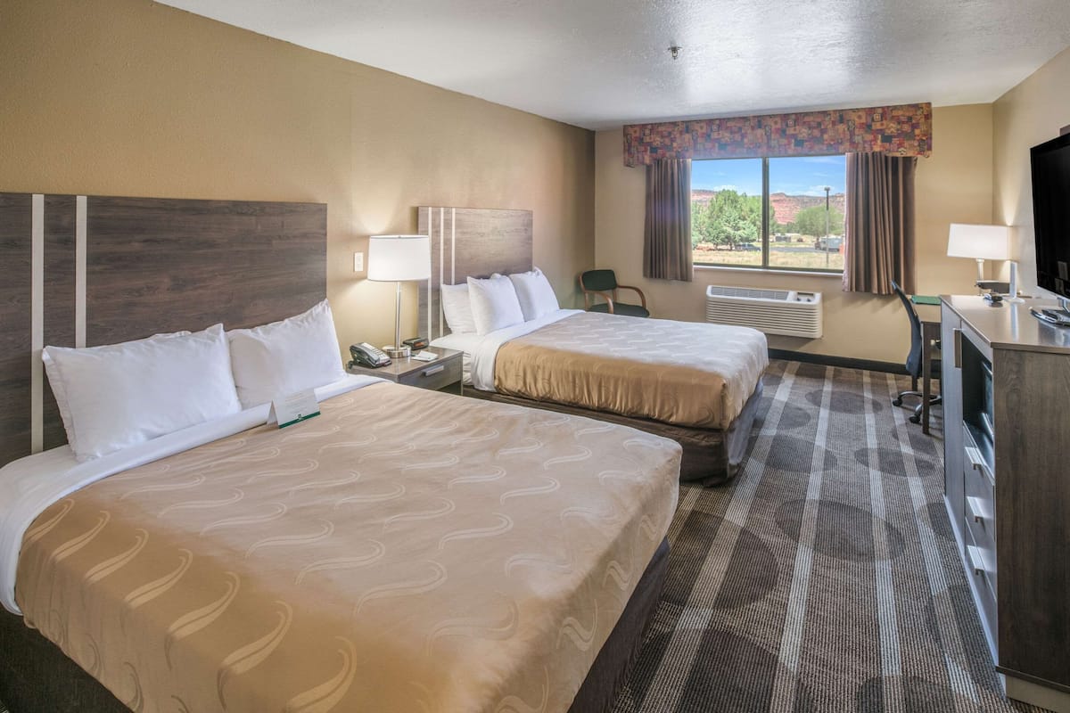 A hotel room with two beds, a desk, a chair, a TV, and a window with a view of the outdoors.