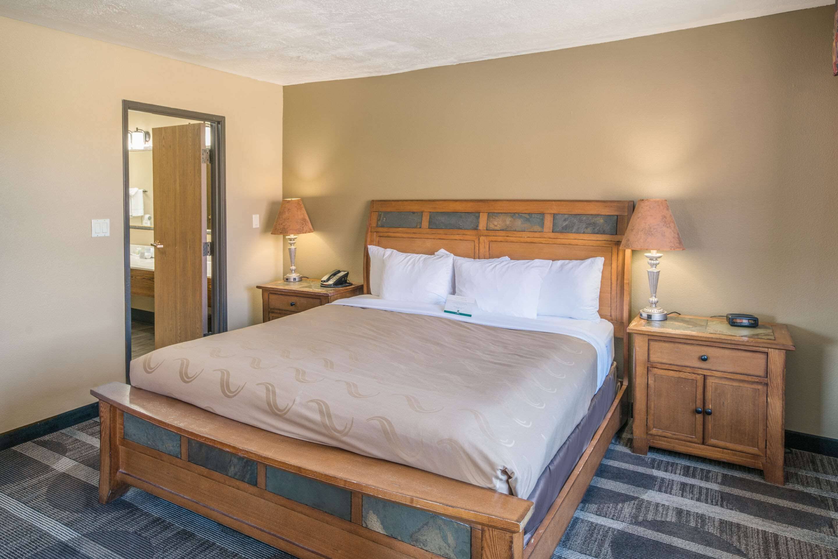 Habitación de hotel con una cama de madera, dos mesitas de noche con lámparas, un espejo y suelo alfombrado.