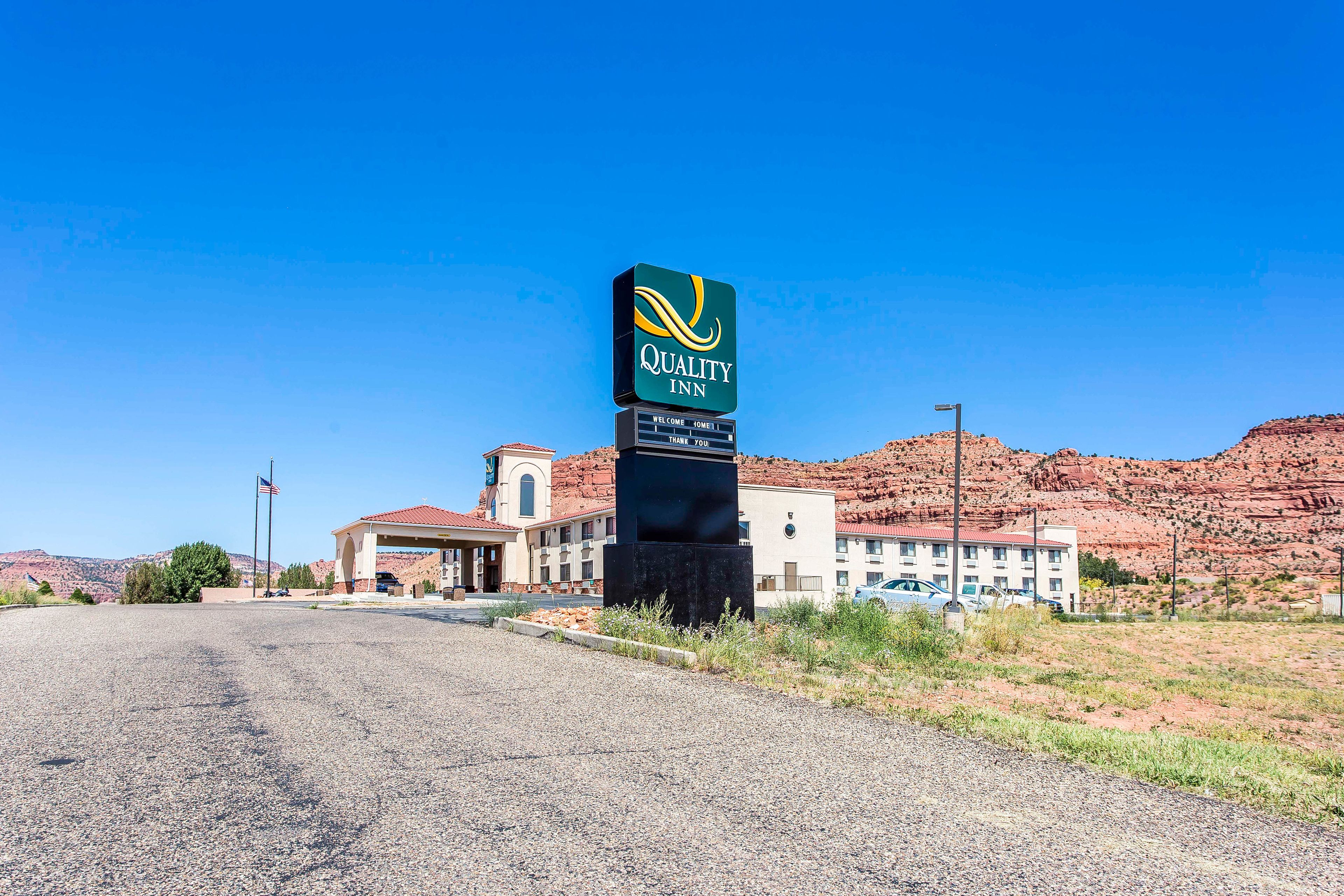 Un letrero de Quality Inn con un mensaje acogedor, un edificio con techo de tejas rojas y montañas al fondo.