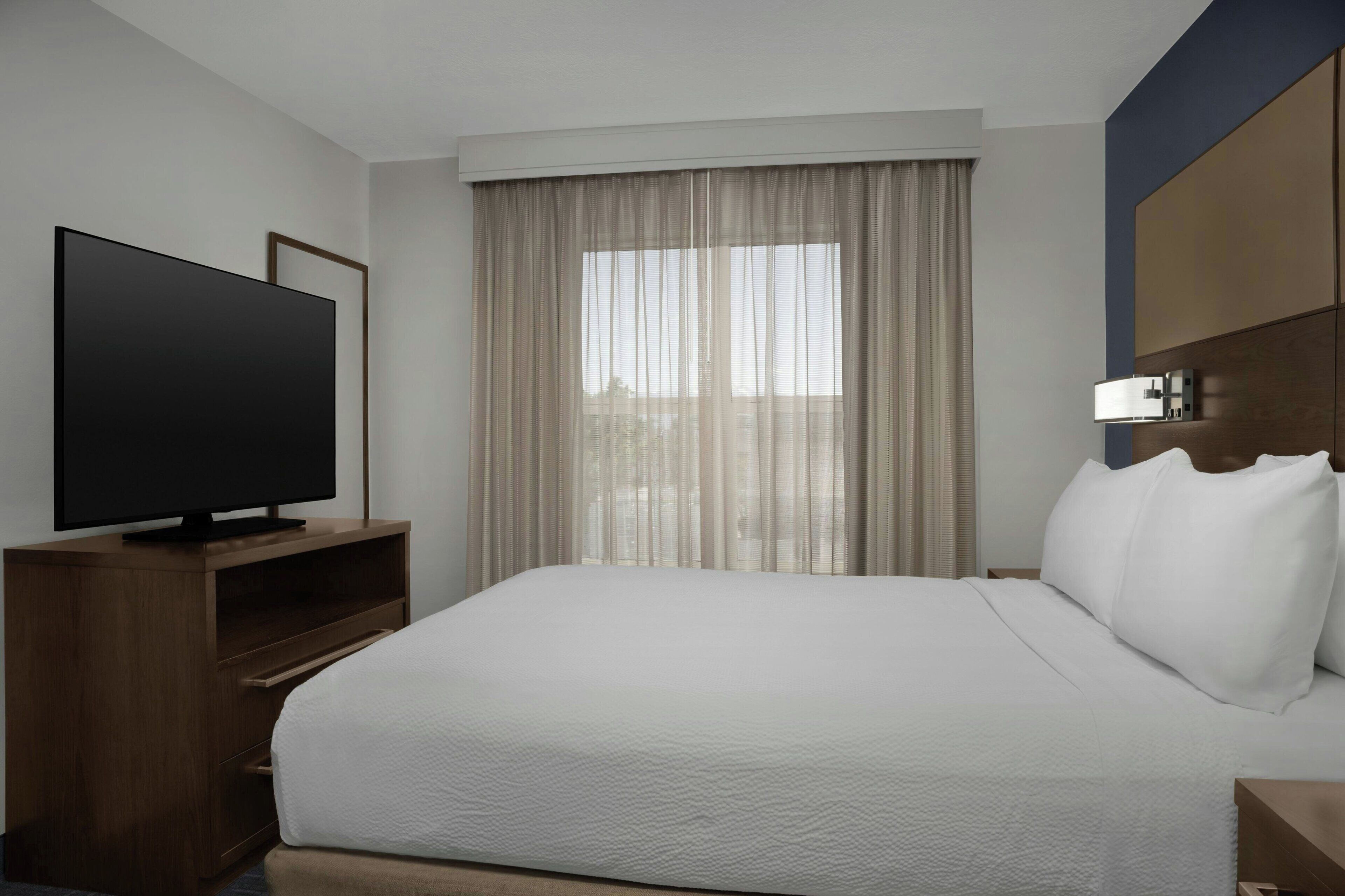 Habitación de hotel con cama, televisor sobre un soporte de madera, ventana con cortinas y cabecera con lámpara.